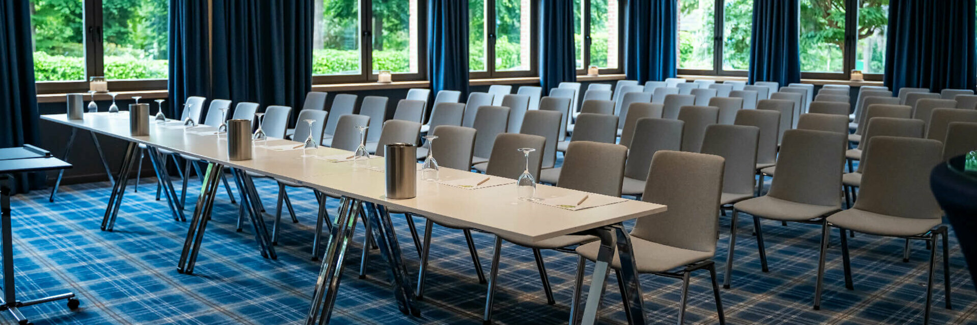 Moderner Konferenzraum im ATLANTIC Hotel mit blauen Vorhängen, Fensterfront, grauen Stühlen und gedecktem Tisch.