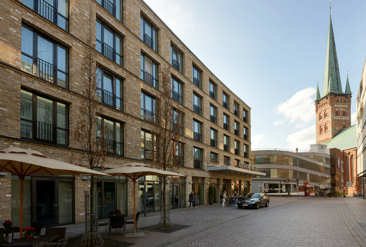 Modern brick hotel building with large windows, outdoor terrace and church tower in the background in sunny weather.