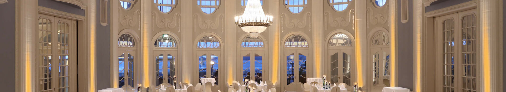 Historischer Ballsaal im ATLANTIC Grand Hotel Travemünde Eleganter Ballsaal mit runden, weiß gedeckten Tischen, hohen Fenstern und großen Kronleuchtern im ATLANTIC Grand Hotel Travemünde.