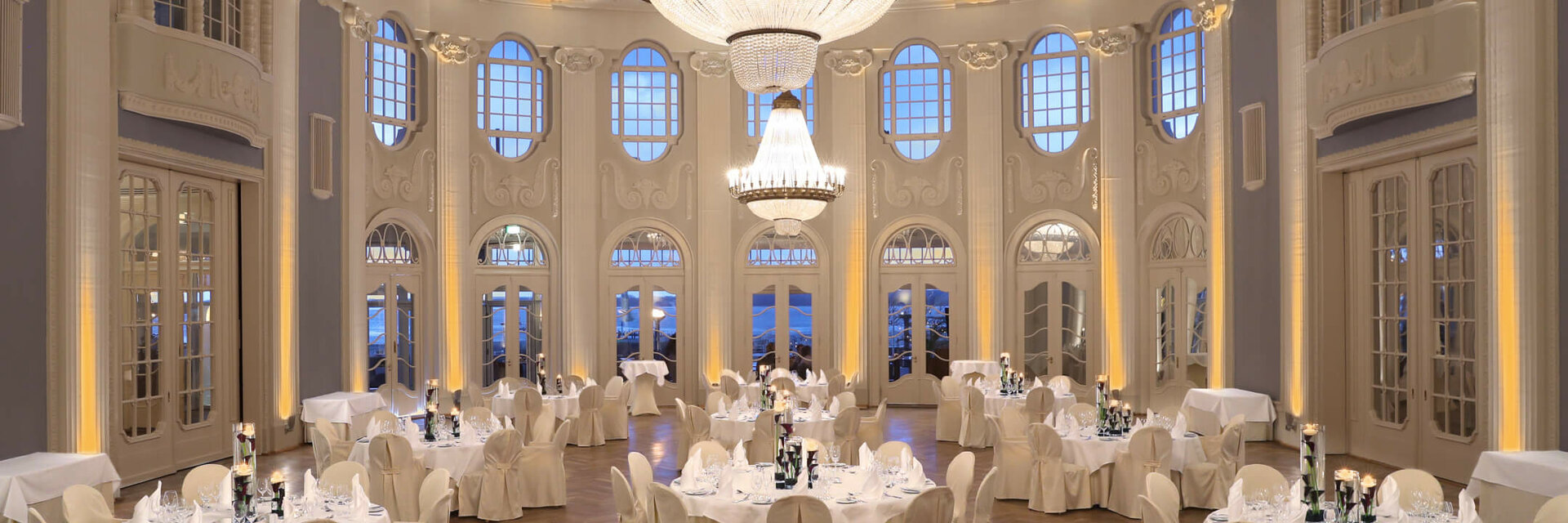 Elegant ballroom with round, white-clothed tables, high windows and large chandeliers in the ATLANTIC Grand Hotel Travemünde.