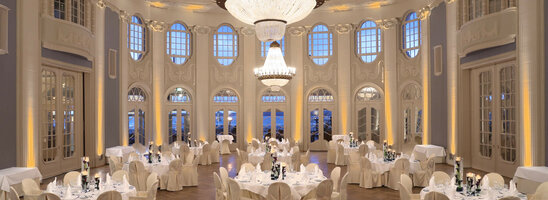 Elegant ballroom with round, white-clothed tables, high windows and large chandeliers in the ATLANTIC Grand Hotel Travemünde.