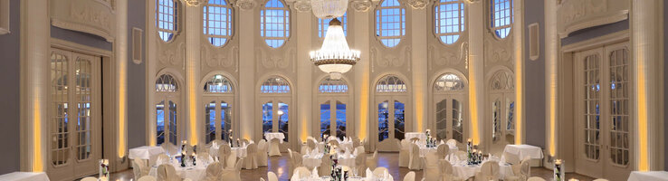Historischer Ballsaal im ATLANTIC Grand Hotel Travemünde Eleganter Ballsaal mit runden, weiß gedeckten Tischen, hohen Fenstern und großen Kronleuchtern im ATLANTIC Grand Hotel Travemünde.