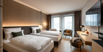 Modernes Hotelzimmer mit zwei Einzelbetten, Schreibtisch, Sessel und großen Fenstern mit Vorhängen im ATLANTIC Hotel Heidelberg.