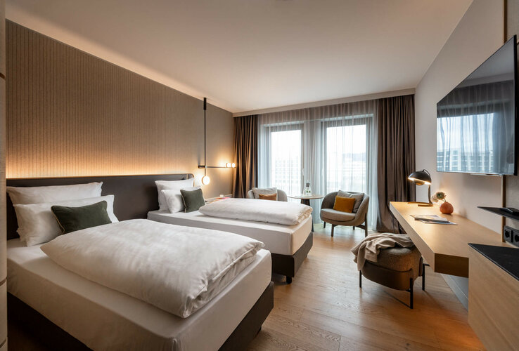 Modern hotel room with two single beds, desk, armchair and large windows with curtains at the ATLANTIC Hotel Heidelberg.