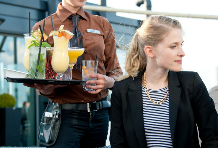 Eine Kellnerin serviert Cocktails auf der Terrasse des ATLANTIC Hotel Kiel. Eine Frau sitzt am Tisch.