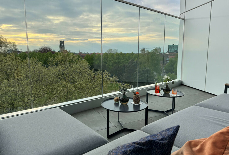 Modern hotel terrace with glass railings, comfortable couch and view of green trees and church tower in the background.