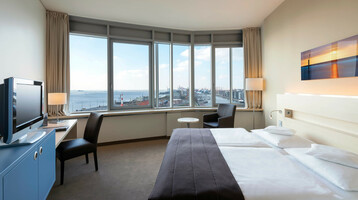 Bright hotel room with double bed, desk, armchair and large window with a view of Bremerhaven harbor.