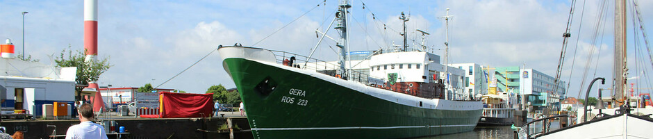 Personen entspannen auf Liegestühlen am Hafen von Bremerhaven, mit Blick auf ein großes, grünes Schiff.