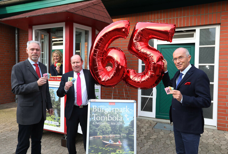 Three men in suits hold red "65" balloons and raffle tickets in front of a kiosk.