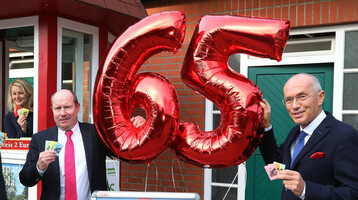 Drei Männer in Anzügen halten rote "65"-Luftballons und Tombola-Lose vor einem Kiosk.