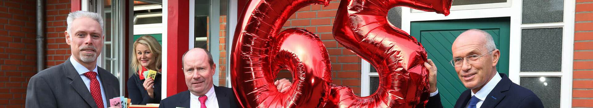 65. Bürgerpark-Tombola Bremen | ATLANTIC Hotels News Three men in suits hold red "65" balloons and raffle tickets in front of a kiosk.