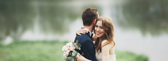 Ein Brautpaar umarmt sich vor einem See im Grünen, die Braut hält einen Blumenstrauß.