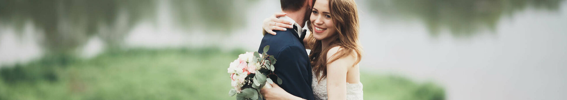 A bride and groom embrace in front of a lake in the countryside, the bride holds a bouquet of flowers.