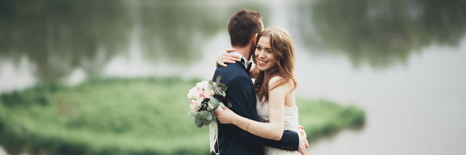 Ein Brautpaar umarmt sich vor einem See im Grünen, die Braut hält einen Blumenstrauß.