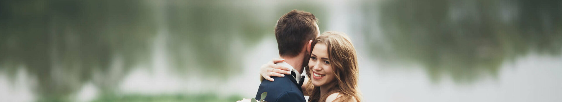 Ein Brautpaar umarmt sich vor einem See im Grünen, die Braut hält einen Blumenstrauß.