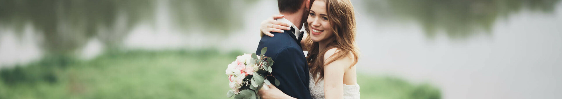 Ein Brautpaar umarmt sich vor einem See im Grünen, die Braut hält einen Blumenstrauß.