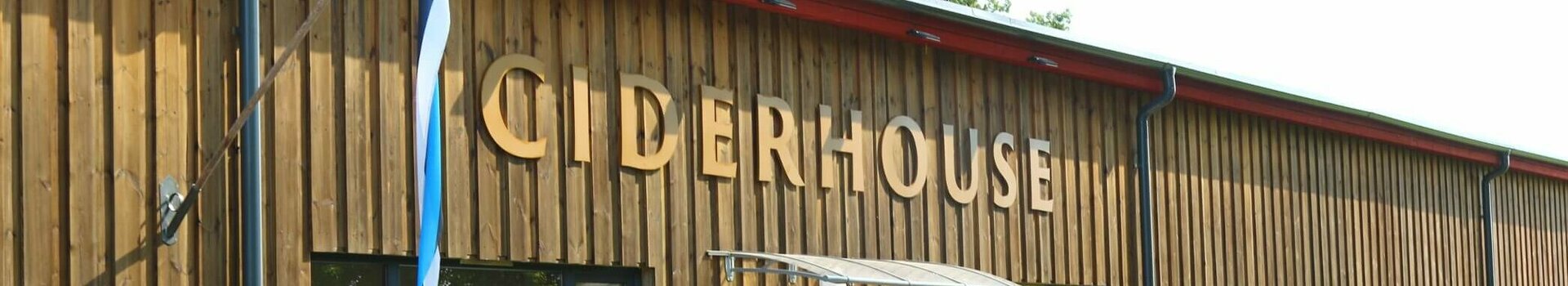 Wooden building with red door, labeled "Ciderhouse", window sills decorated with flowers and Estonian flag.