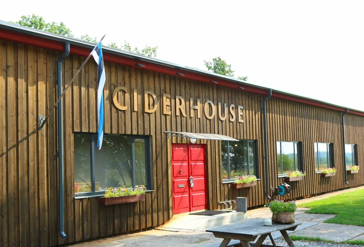 Wooden building with red door, labeled "Ciderhouse", window sills decorated with flowers and Estonian flag.