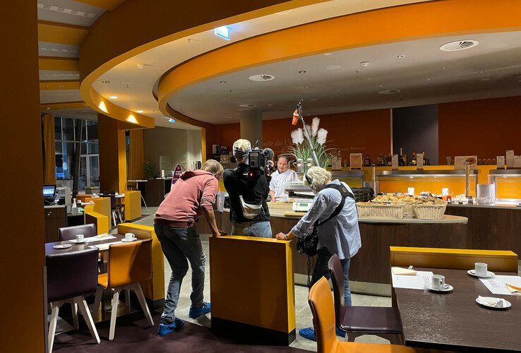 Hotel restaurant with breakfast buffet; camera team films chef behind buffet, modern furnishings in warm colors.