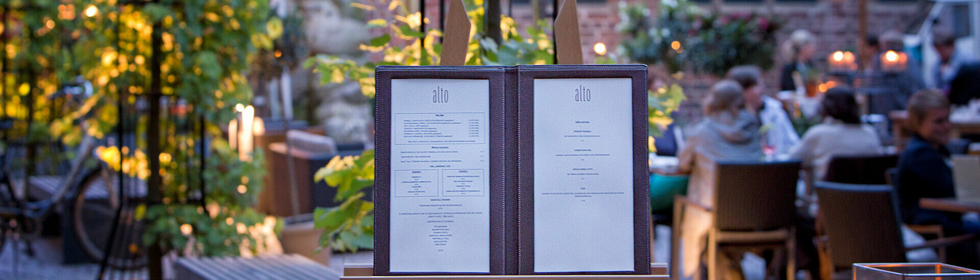Restaurant menu on an easel, surrounded by an illuminated, green inner courtyard with guests at the ATLANTIC Grand Hotel Bremen.