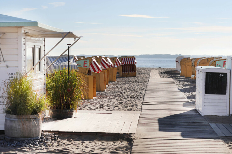 Holzsteg führt zu Strandkörben am Sandstrand, Meer im Hintergrund, links weiße Hütte mit Pflanzen.
