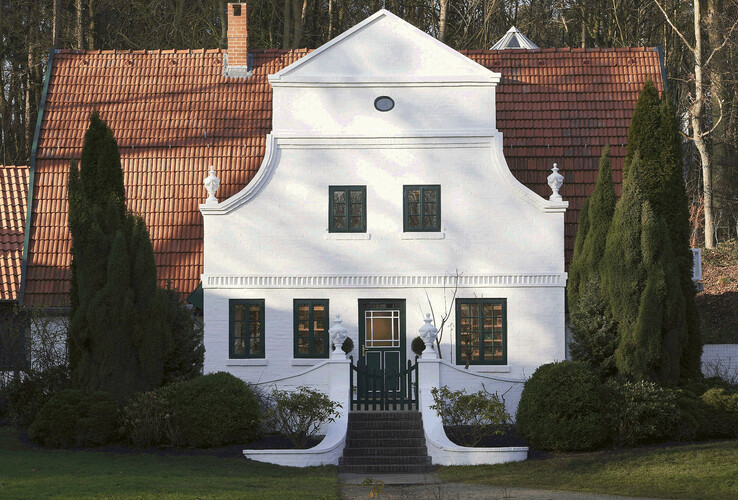 Weißes, historisches Gebäude mit rotem Ziegeldach, umgeben von Bäumen und gepflegtem Garten.