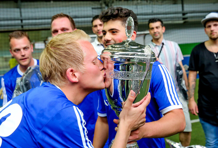 Ein Fußballspieler in blauem Trikot küsst einen großen silbernen Pokal, umgeben von jubelnden Teamkameraden.
