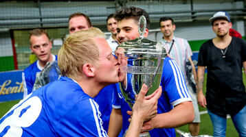 A soccer player in a blue jersey kisses a large silver trophy, surrounded by cheering teammates.
