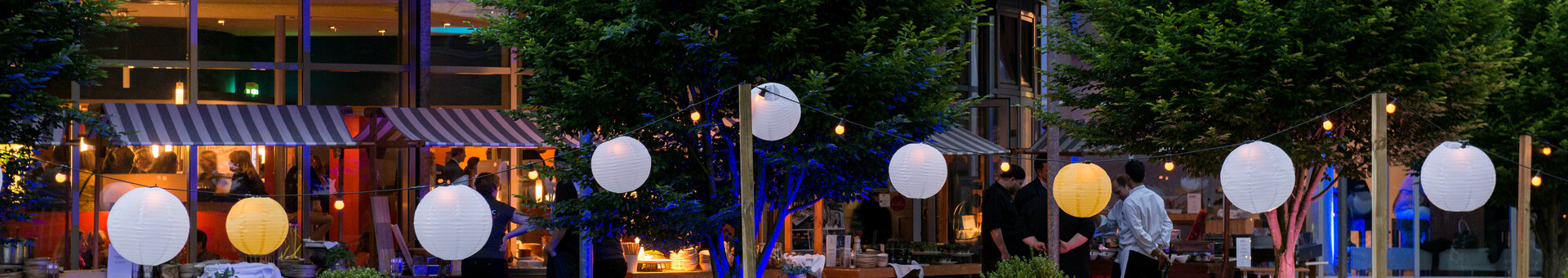 Terrasse des ATLANTIC Hotel Universum in Bremen Außenbereich des ATLANTIC Hotel Universum mit beleuchteten Lampions, Tischen und Stühlen, umgeben von Bäumen.