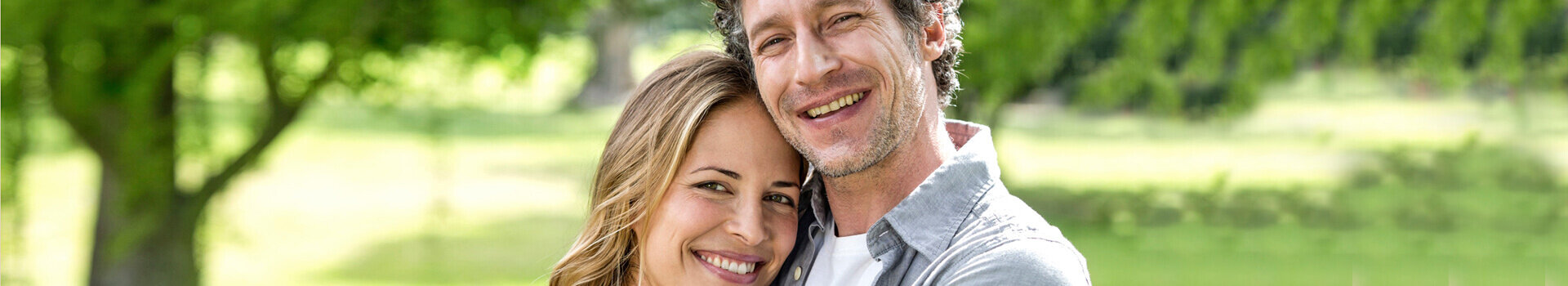 Ein lächelndes Paar steht umarmt in einem grünen Park, Bäume im Hintergrund.