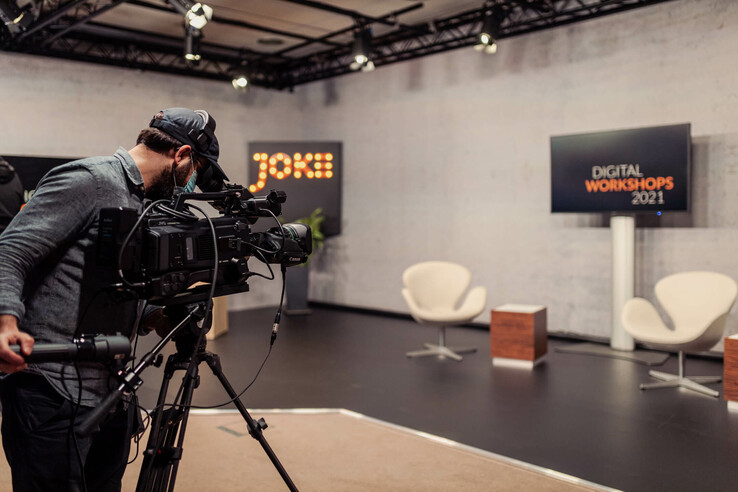 Ein Kameramann filmt ein modernes Studio mit zwei weißen Sesseln und einem Bildschirm für digitale Workshops.