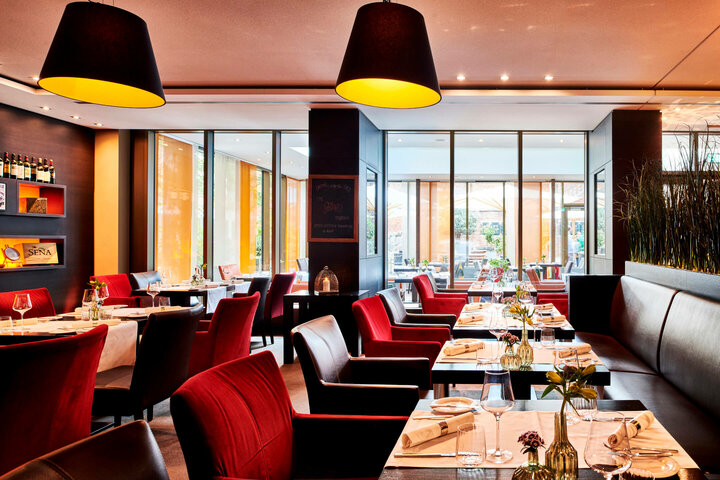 Restaurant alto ATLANTIC Grand Hotel Bremen Modernes Hotelrestaurant mit roten Stühlen, gedeckten Tischen und großen Fenstern mit Blick auf die Terrasse.
