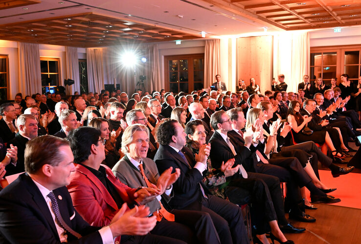 Eine große Gruppe von Menschen in formeller Kleidung applaudiert in einem elegant beleuchteten Veranstaltungsraum.