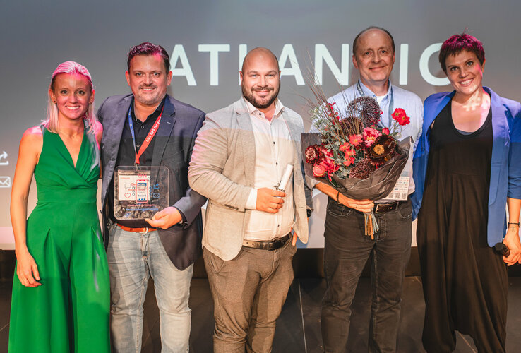 Fünf Personen lächeln auf einer Bühne, eine hält Blumen, eine andere eine Auszeichnung. Hintergrund: ATLANTIC Hotel.