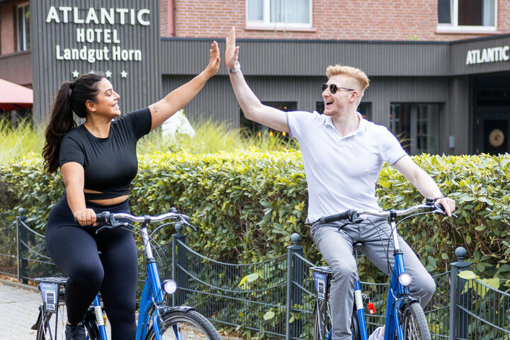 Zwei Personen auf Fahrrädern geben sich vor dem ATLANTIC Hotel Landgut Horn fröhlich ein High-Five.