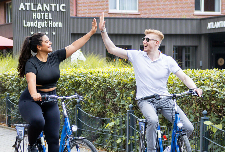 Zwei Personen auf Fahrrädern geben sich vor dem ATLANTIC Hotel Landgut Horn fröhlich ein High-Five.