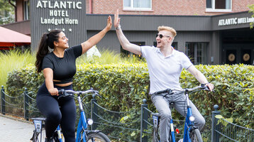 Two people on bicycles give each other a cheerful high-five in front of the ATLANTIC Hotel Landgut Horn.