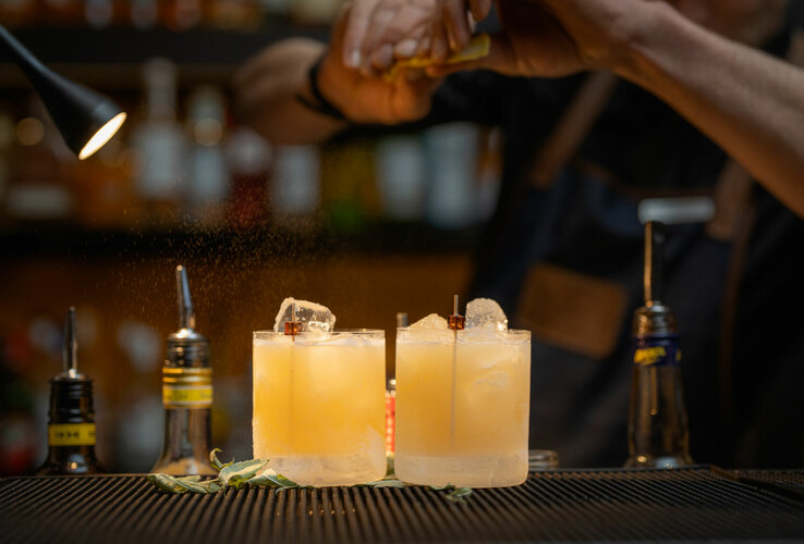 Zwei Cocktails auf einer Bar, mit Eiswürfeln und dekorativen Zutaten, werden von einem Barkeeper zubereitet.