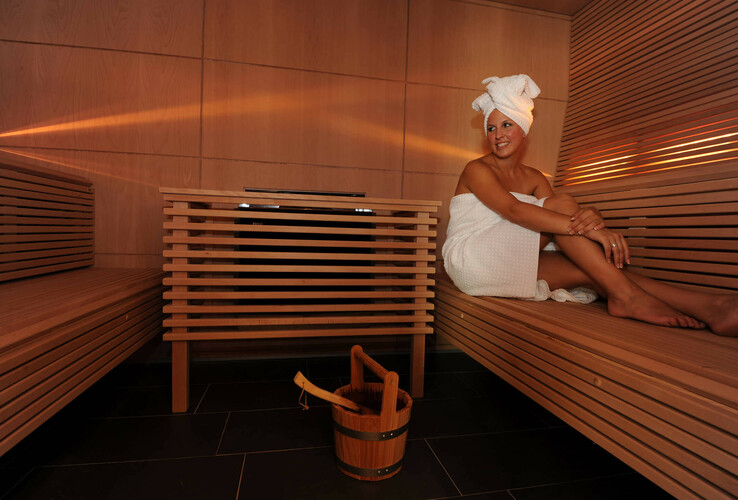 Frau in Sauna des ATLANTIC Grand Hotel Bremen, eingewickelt in ein Handtuch, entspannt auf Holzbank sitzend. 