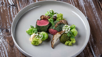 Gourmet dish with pink roasted meat, green herbs, mushrooms and floral decoration on a gray plate.