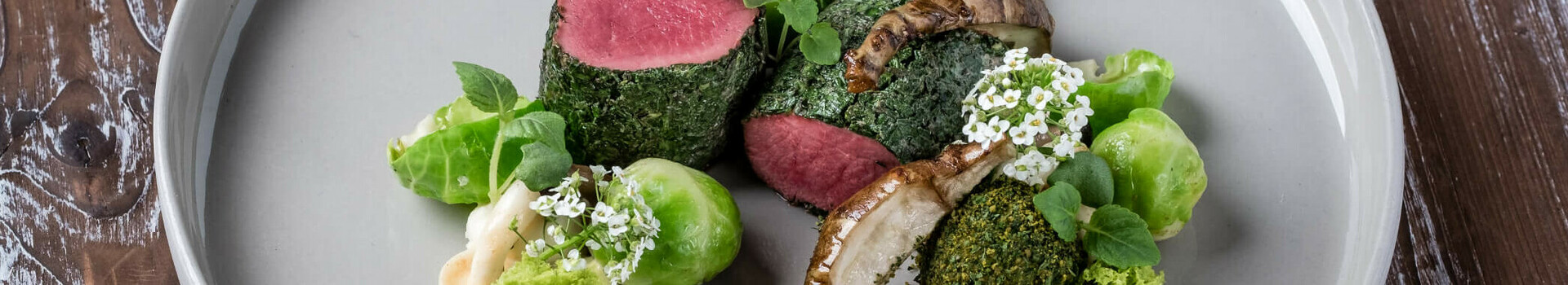 Gourmetgericht mit rosa gebratenem Fleisch, grünen Kräutern, Pilzen und Blumendekor auf grauem Teller.