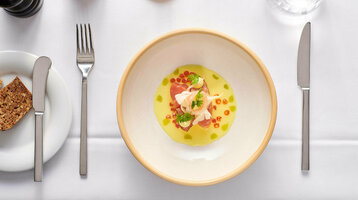 Gourmet-Vorspeise in stilvoller Anrichtung auf weißem Tisch, mit Brot, Besteck und einem Glas Wasser.