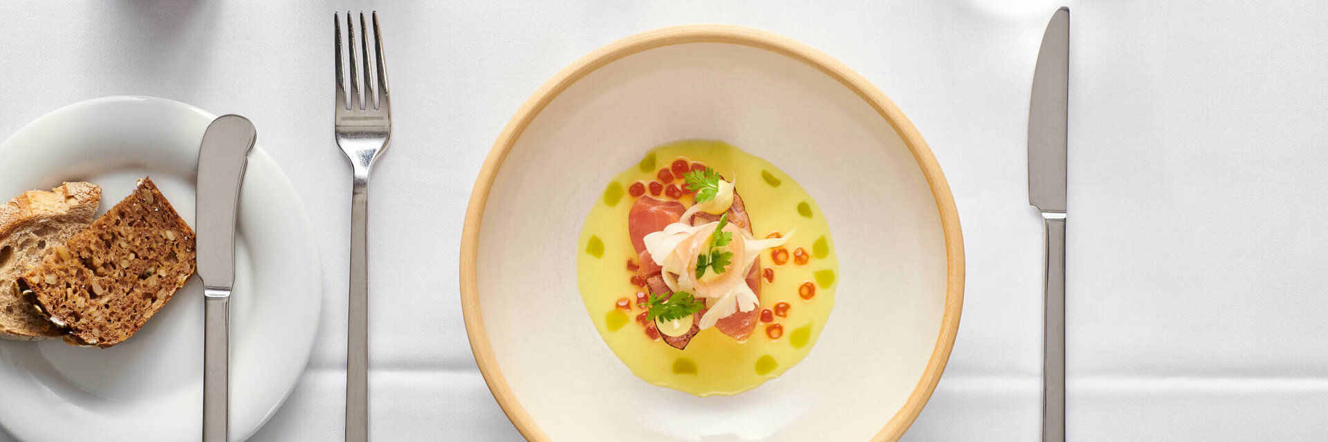 Gourmet-Vorspeise in stilvoller Anrichtung auf weißem Tisch, mit Brot, Besteck und einem Glas Wasser.