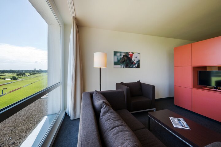 Platz, Stil und Komfort in unserem Studio mit Blick auf die Rennbahn Modernes Hotelzimmer mit braunem Sofa, Sessel, Lampe und Blick auf die Galopprennbahn durch ein großes Fenster.