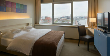 Bright hotel room with double bed, view of the harbor through large windows, TV and desk with lamp.
