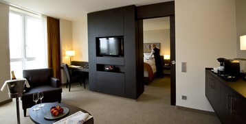 Modern hotel room with sitting area, desk, TV, bed in the background and view of a person making the bed.