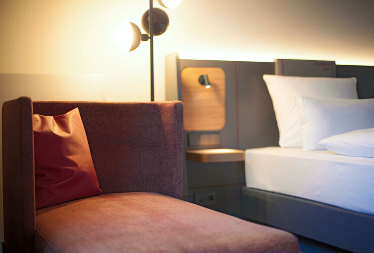 Business Zimmer im ATLANTIC Hotel Airport Bremen Modernes Hotelzimmer mit gemütlicher Sitzecke, beleuchtetem Nachttisch und einem Bett mit weißen Kissen im ATLANTIC Hotel.