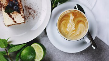 Kaffee mit Latte Art und Dessert auf einem Tisch, daneben Limetten und Minzblätter.