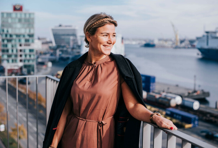 Woman on hotel balcony with view of Kiel harbor, smiling, in sunny weather.