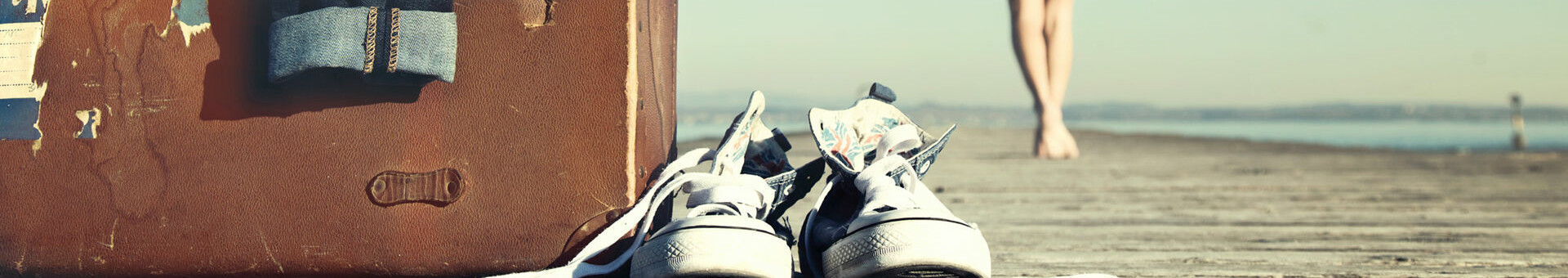 Alter Koffer und Sneakers auf einem Steg, Person in Bademantel geht zum Wasser.